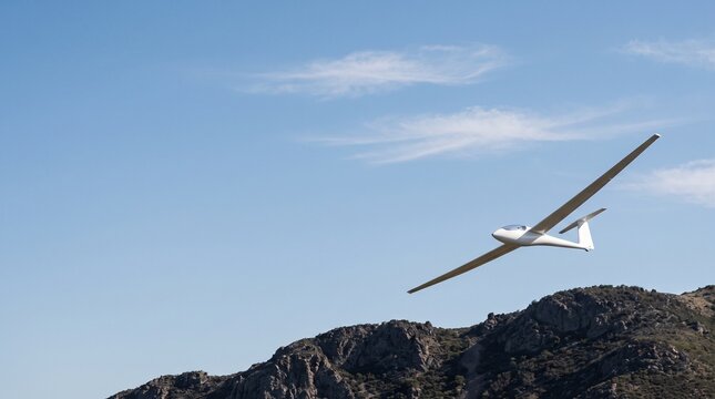 A white glider airplane soars gracefully over a rugged, rocky mountain landscape under a clear blue sky with wispy clouds.