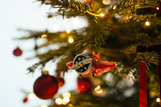 Close up vertical shot of a retro toy plane bauble decorating a green Christmas tree branch with warm glowing holiday fairy lights in the background.
