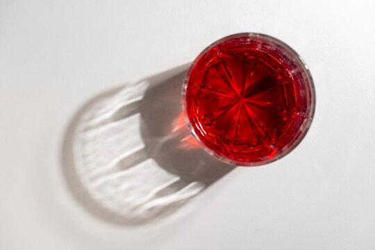 Top view of red drink in crystal glass with beautiful shadow, closeup. High-angle shot of faceted glass filled with vibrant red liquid, casting patterned, caustic shadow on white background