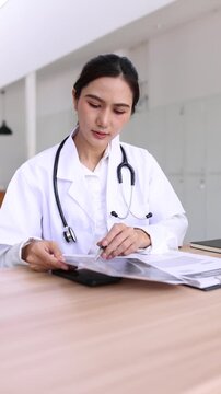 Asian female doctor using digital tablet for online video call consultation with patient and reading medical report in hospital office.