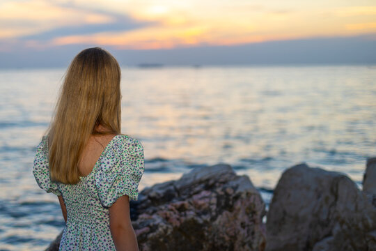 Young Girl Looking at the Sea at Sunset. Young girl in a floral dress standing by rocky shore and looking across the sea at sunset.