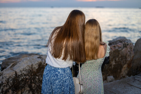 Sisters Looking at the Sea at Sunset. Two sisters standing together on a rocky shore and looking across the calm sea at sunset.