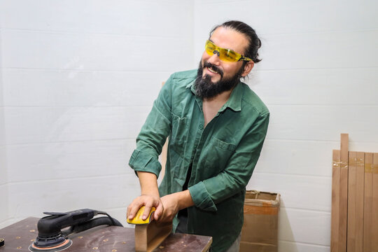 SMILING CARPENTER SANDING CURVED WOOD PIECE AT WORKSHOP BENCH WITH HANDHELD SANDER, WEARING YELLOW SAFETY GLASSES AND GREEN WORK SHIRT, PREPARING SURFACE FOR SMOOTH FINISH