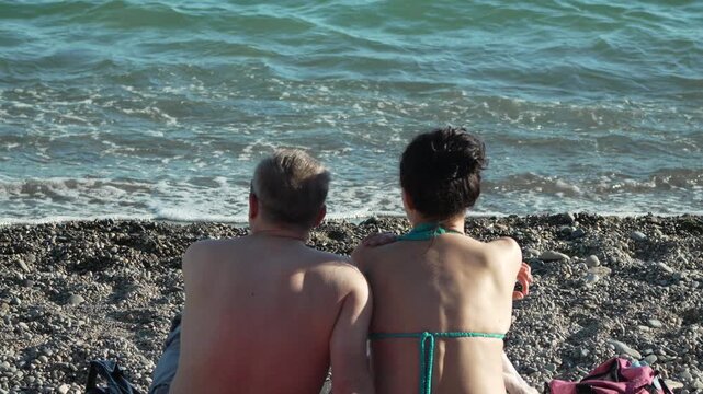 Couple relaxing together on rocky beach while viewing calm turquoise ocean waves. Static medium shot from a rear perspective.