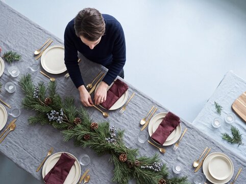 High angle view of a man setting a Christmas holiday dinner table decorated with pine garland and golden cutlery