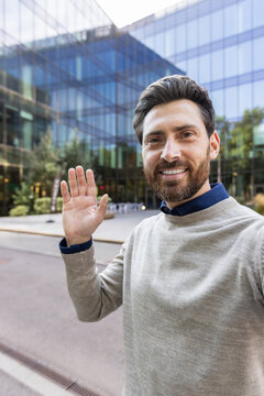 Vertical close-up photo of a young smiling man standing outside, holding a camera, talking, smiling and waving