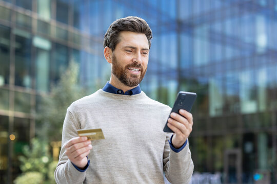 Smiling young man standing on the street, holding a credit card in his hand and using his mobile phone