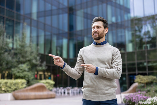 Smiling young man standing outside, pointing with fingers at empty background and looking to the side
