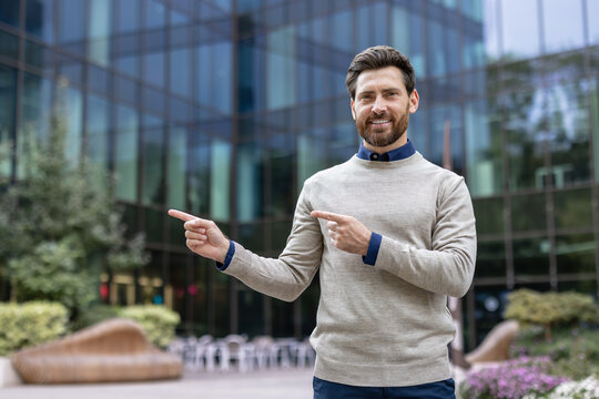 Portrait of a young man standing outside on the street and pointing with his fingers at an empty space next to him, looking at the camera with a smile