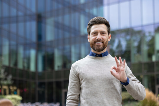 Portrait of a successful young man standing outside an office building, looking at the camera and showing the OK sign with his hand