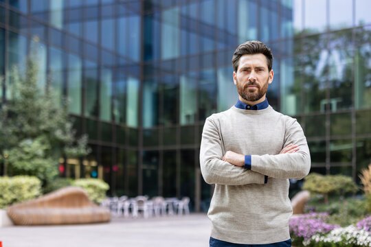 A serious young businessman is standing near an office center on the street, arms crossed on his chest and looking confidently at the camera