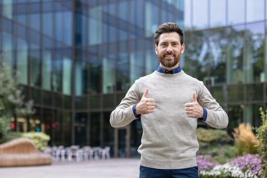 Portrait of a young happy man standing on the street outside an office center, looking at the camera and showing super signs with his fingers