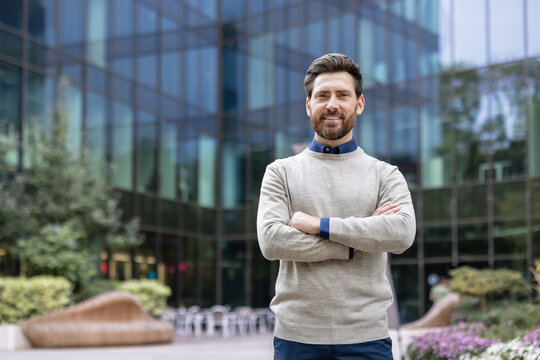 Smiling young businessman standing near an office center on the street, arms crossed on his chest and looking confidently at the camera