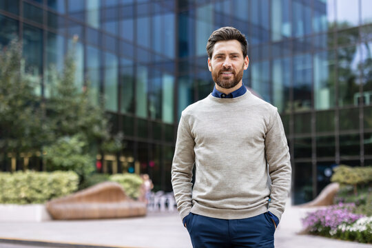 Portrait of a young successful businessman standing outside an office building, holding his hands in his pockets and looking at the camera with a smile