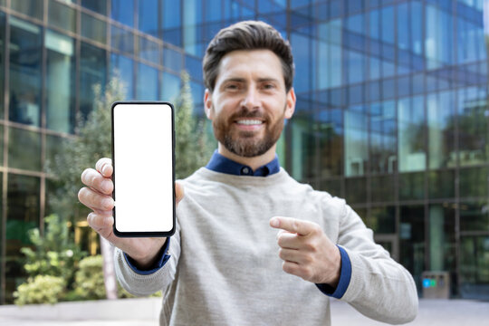 Portrait of a young smiling man standing outside and pointing with his finger at the camera a mockup phone with a white screen