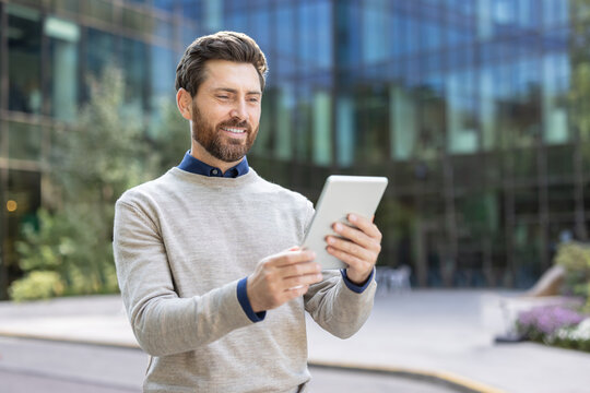 A smiling young man is standing on the street and looking at the screen of the tablet he is holding in his hands