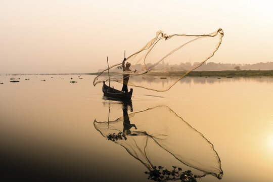 Fisherman with boat on Taung Tha Man Lake casts a fishing net for sunrise, reflection in the water, Thaung Tha Man Lake, Mandalay, Myanmar