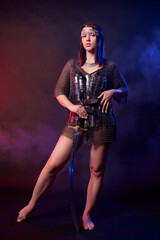 Fototapeta premium A portrait of a young woman in oriental chainmail armor, holding a saber and posing standing against a dark background with red and blue backlighting. Concept art of an oriental warrior.