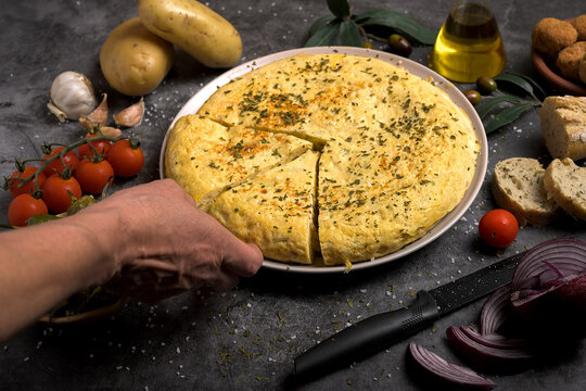 Hand taking a portion of juicy traditional Spanish omelette on a plate with bread, oil, tomatoes and peppers on dark background