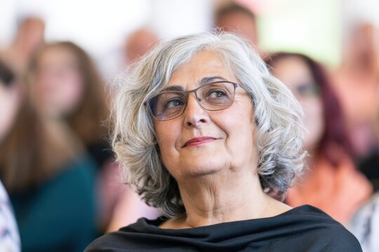 Attractive woman in her early 60s with short wavy gray hair and glasses attentively listens during a community lecture, surrounded by a softly blurred audience