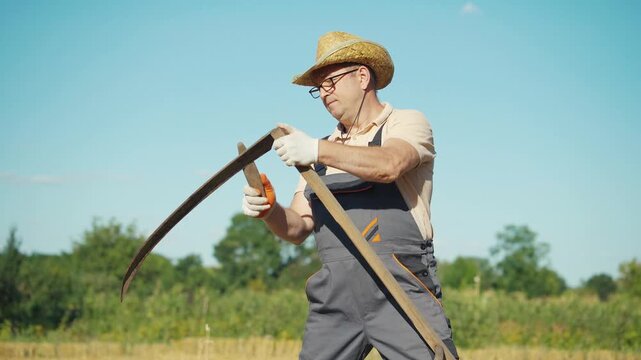 Older man in straw hat and gloves sharpens a scythe while standing in a sunny field, surrounded by greenery and blue sky, preparing for agricultural work in a rural setting