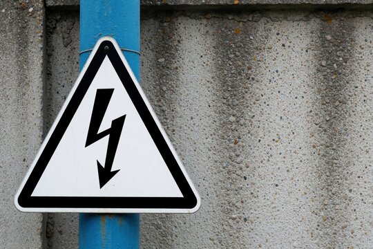 Triangular high voltage warning sign affixed to a blue pole against a textured concrete wall. Symbol indicates danger of electrical shock and imminent hazard.