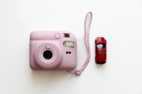 A pink instant camera is placed beside a small red toy car on white background, creating a visually appealing composition that highlights both objects in a bright and minimalistic setting
