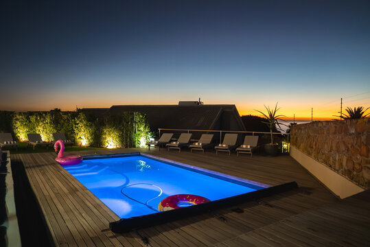 An illuminated swimming pool rests on a timber deck with sun loungers and floats during evening sunset twilight in Cape Town, South Africa.