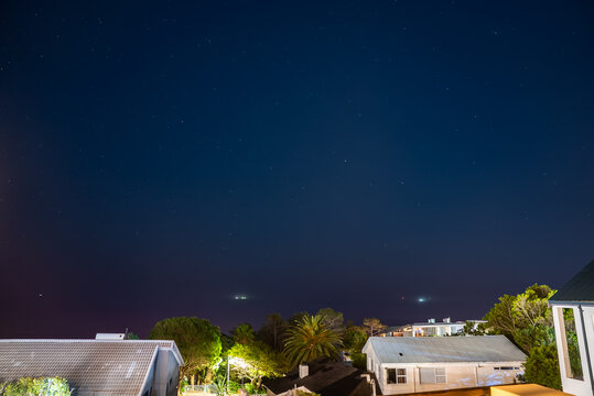 A clear dark night sky filled with visible stars extends over suburban rooftops, palm trees, and illuminated gardens in Cape Town, South Africa.