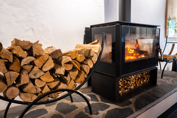 Fototapeta premium A contemporary black glass wood stove burns brightly beside a metal cradle filled with cut firewood logs sitting on a stone floor inside a living space.