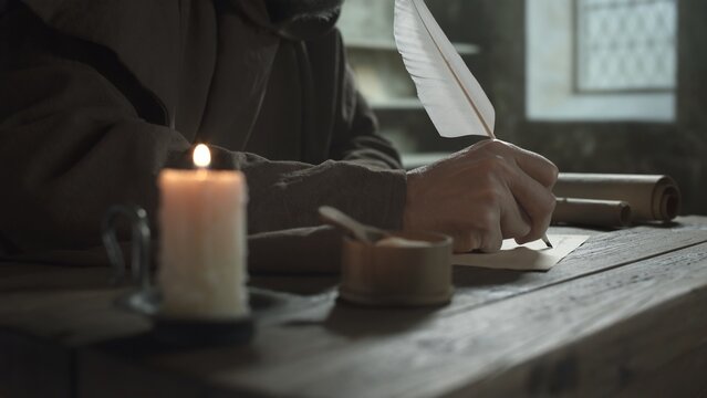 Hands of a monk writing with a large quill pen on parchment on a rustic table near a burning candle inside a medieval monastery library room