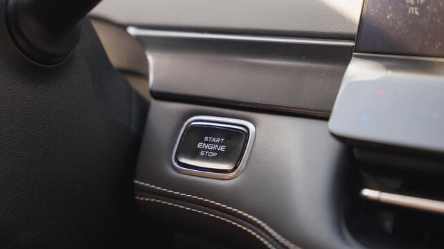 Soft focus hand with ring presses start engine button inside modern car cabin, framed by dark textured trim, glossy panel edge, metallic accent strip and stitched leather under warm natural light