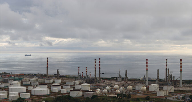Portugal - 15 August 2026: Aerial view of an oil refinery with storage tanks and tall chimneys along the coast, with a cargo ship on the ocean under a cloudy sky.