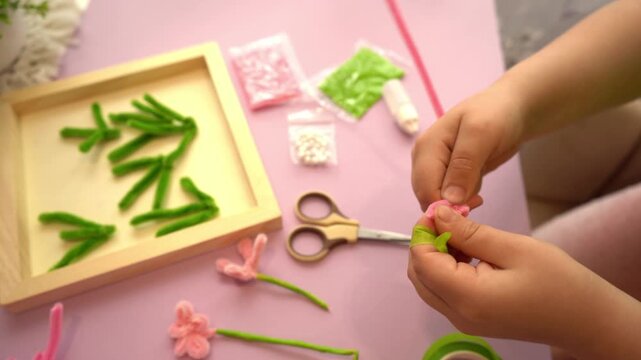 Unrecognizable child crafting a colorful fuzzy wire flower using bright pink and green chenille sticks on a purple desk backdrop during a creative home lesson or hobby class