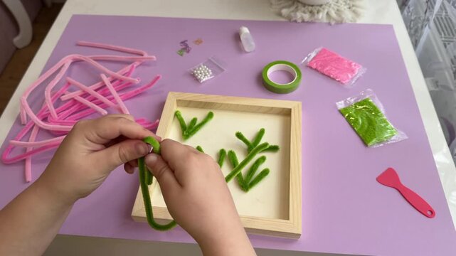 Top view of an intelligent child working with a craft kit twisting pink pipe cleaners over a wooden workspace frame for cognitive skills enhancement advertisement