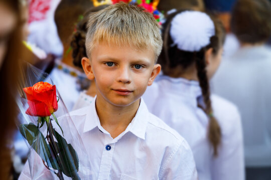 A young schoolboy in a crisp white shirt holds a fresh red rose during a formal outdoor assembly celebrating the start of a new autumn school year today