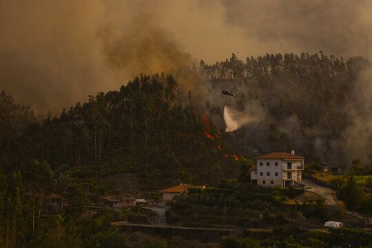 Arouca, Portugal - 20 August 2026: View of a firefighting helicopter dropping water on a forest fire burning on a hillside near a house in the rural landscape.