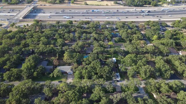 Concrete highway barriers buffer quiet single story residential homes along Airport Freeway. Towering live oaks, hidden asphalt cul de sacs, detached garages display suburban layout, Hurst, Texas