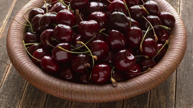 Sweet cherries with stems on a handmade ceramic plate. Fresh organic fruits on a vintage ribbed dish with a weathered, dark rustic wood background, table spin, backlit natural light.