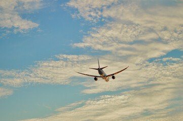 Obraz premium Commercial passenger airplane flying through partly cloudy blue sky with warm golden light illuminating the wings during approach