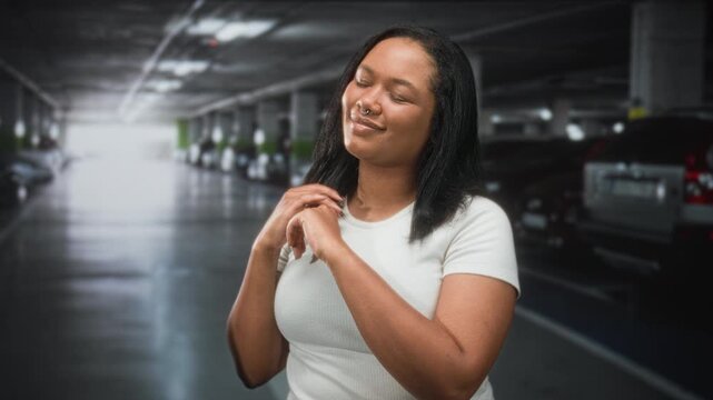 Woman clasping hands near chest, septum ring visible, wearing white tshirt, smiling in parking building; quiet contentment.