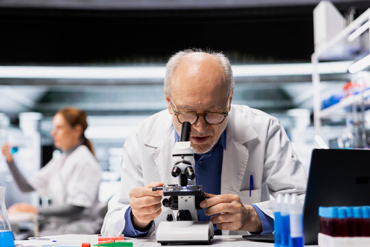 Senior molecular biologist inspects a new drug compound under a microscope for consistency; elderly man in the research facility verifies medication quality and purity standards during analysis.