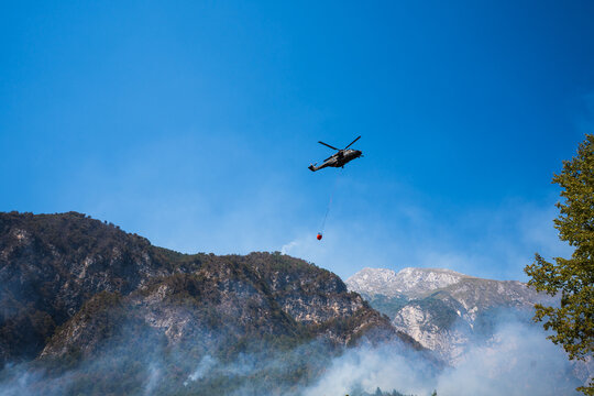 Fire-fighting helicopter fighting a large forest fire on a rocky slope, characterised by thick smoke and high-altitude water drops