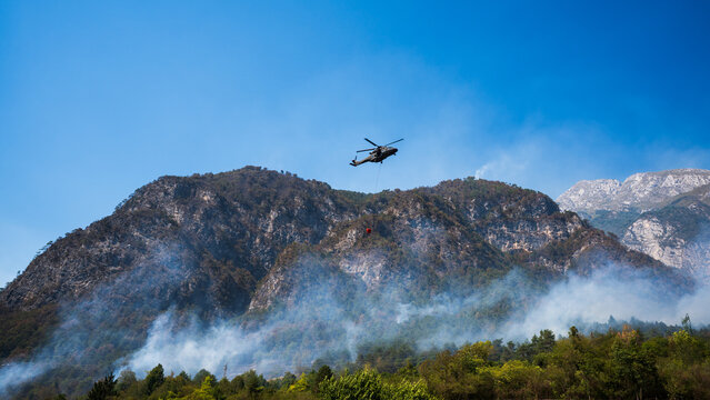 Fire-fighting helicopter fighting a large forest fire on a rocky slope, characterised by thick smoke and high-altitude water drops