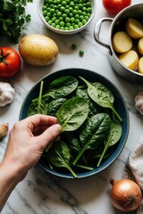 Naklejka premium Hand selects fresh spinach from bowl. Ingredients for cooking include potatoes, peas, tomatoes, garlic, and onion on marble surface. Preparing healthy meal, fresh produce for dinner.