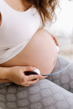 Pregnant woman holding headphones to her belly, playing music for unborn baby at home, maternity and prenatal care concept.