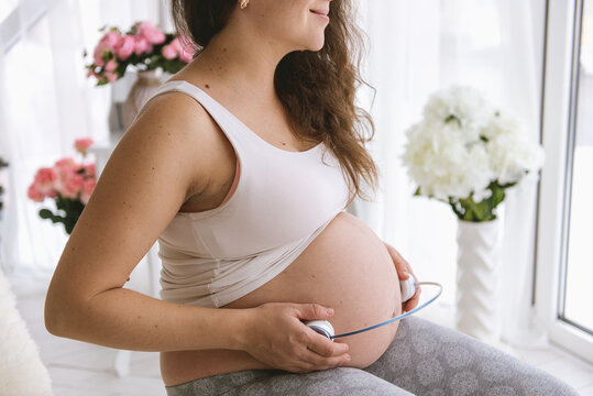 Pregnant woman holding headphones to her belly, playing music for unborn baby at home, maternity and prenatal care concept.