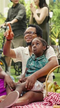 Portrait format: Joyful parents laugh with a boy seated on his father's lap in a city park, enjoying playful conversation on a picnic blanket beside a fruit and pastry basket.