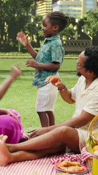Vertical aspect: Boy plays a hand gesture game with parents seated on a picnic blanket beside a food basket in a sunny urban park with skyline views, as the family giggles and enjoys bonding outdoors.