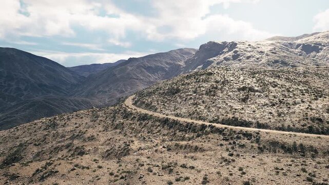 A winding road curves through rugged mountains surrounded by sparse vegetation and rocky terrain. The sky is partly cloudy, creating a dramatic backdrop for the landscape.